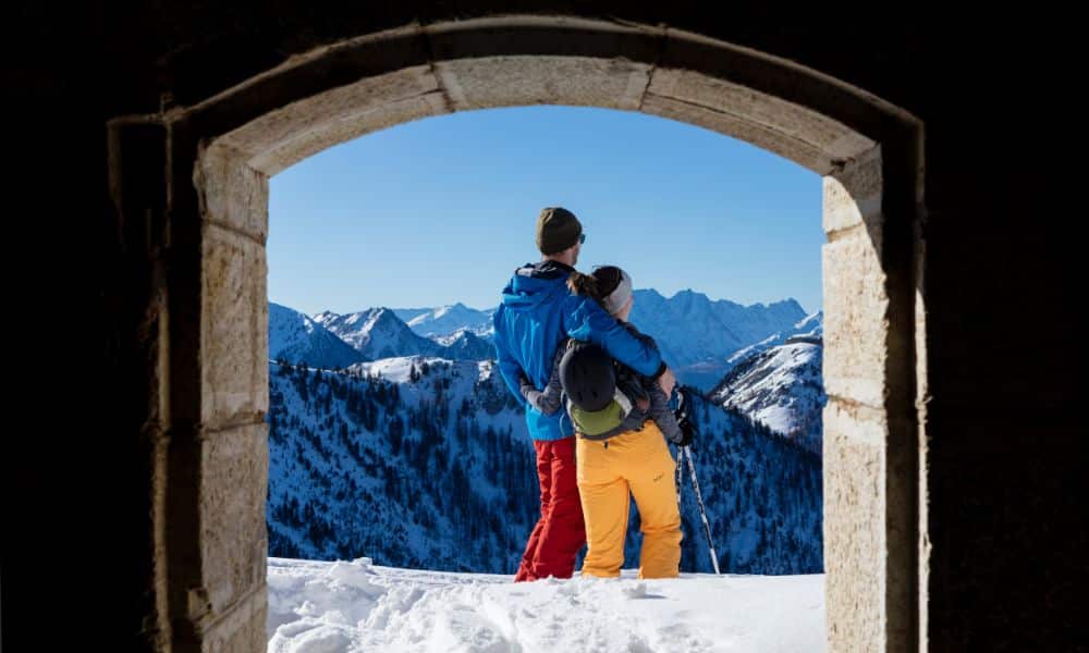 A couple stand together looking at the landscape before them. They are wearing cloths fit for skiing, and also have ski equipment on them 