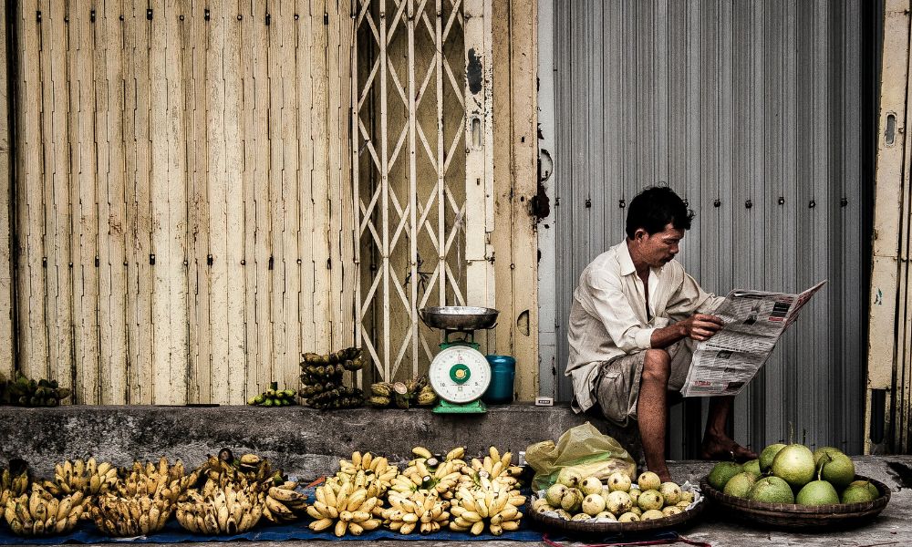 A vendor in Vietnam sits by the road side with his wares on display - bananas and other fruits. Behind him is a weighing scale, the shutters of a shop and he reads a local newspaper 